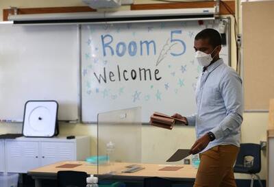 A Black teacher wearing a mask passes out folders onto desks as they ready for the students' arrivals.