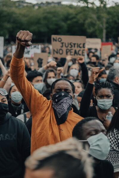 A Black man in an orange shirt holds his fist up in the symbol for unity and solidarity at a protest held in George Floyd's honor. A sign behind the man reads "No Justice, No Peace."