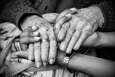 The weathered hands of an elder are held by the hands of a younger person. Their hands rest on a cloth bag.