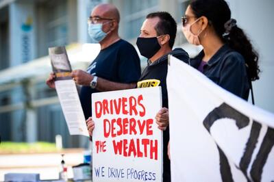 Hector Castellanos (center) with We Drive Progress holds a sign that says 'Drivers Deserve the Wealth' outside DoorDash headquarters in San Francisco, along with fellow gig workers (rideshare and delivery drivers) demanding fair pay and employee rights.